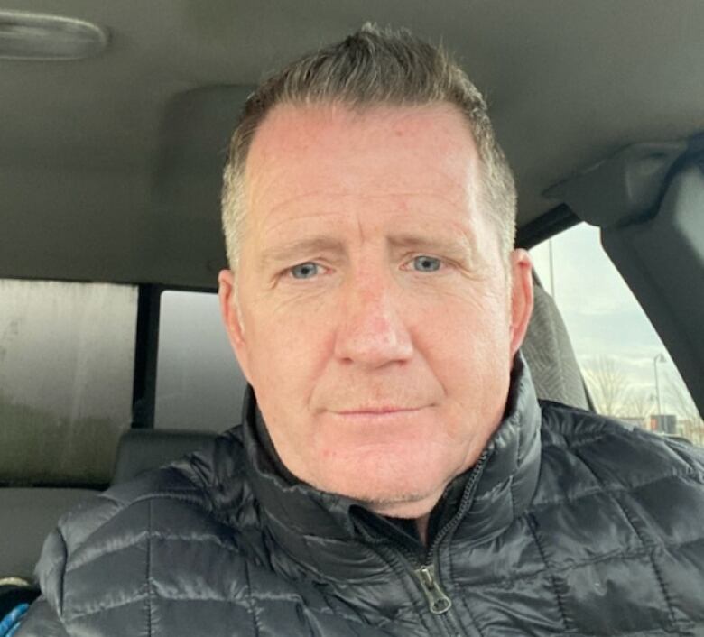 A selfie image of a middle-aged man with reddish, grey hair, sitting in his car.