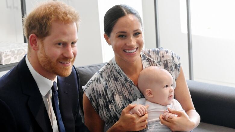  A man with red hair and dressed in a suit next to a woman with long dark hair holding a baby on her lap. 