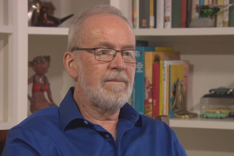  A bearded man in a blue shirt sits in front of a bookcase.