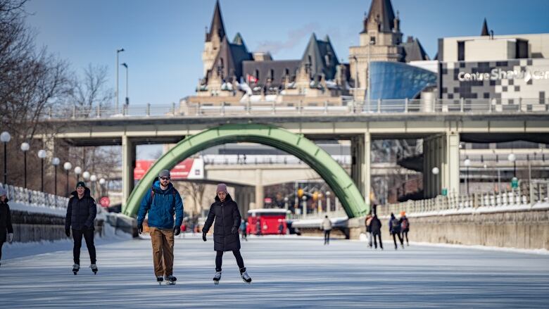 Two people skate under a bridge with a castle-like building in the background.