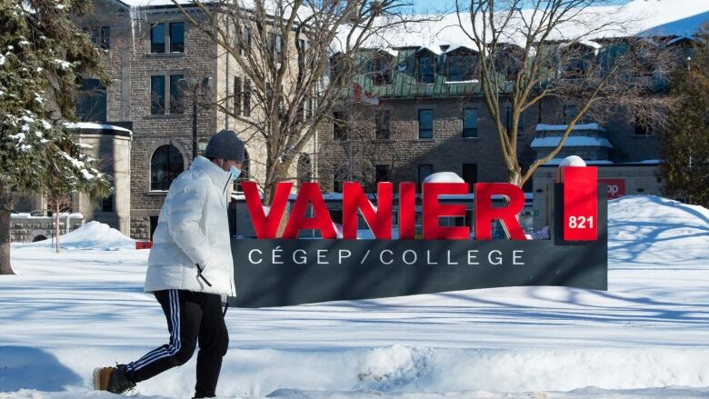 A person in a mask walks past a sign that says Vanier College.