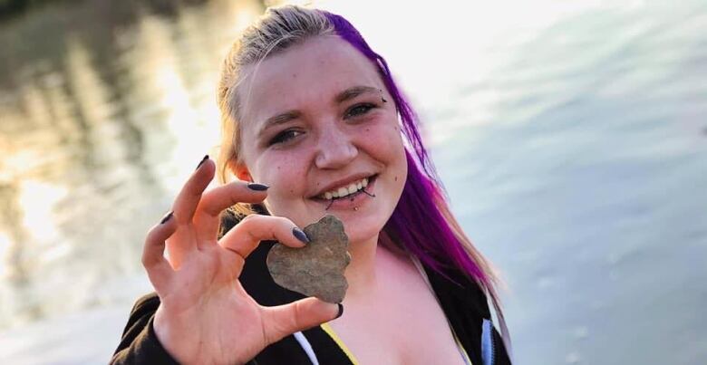 A woman with purple hair holds up a rock near a water body.