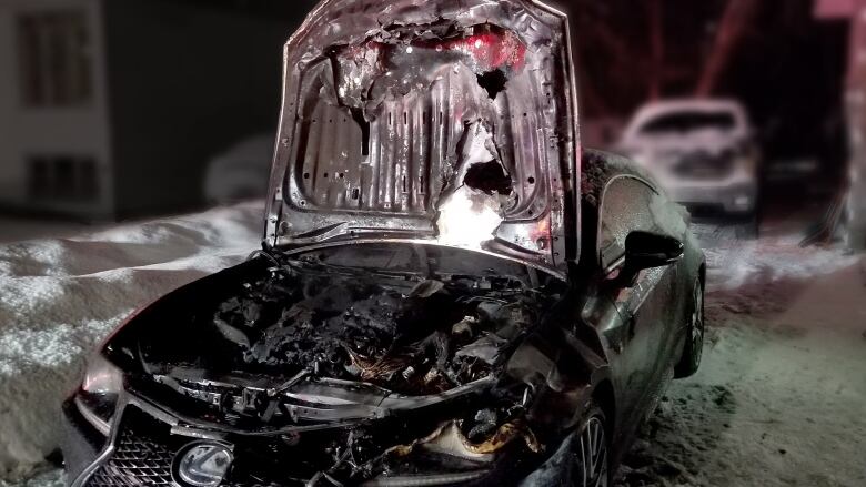 A car parked outdoors at night with snow in the background has its hood up, revealing fire has burned the engine area.