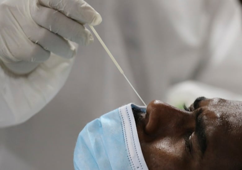 A health-care worker wearing a white medical gown and white gloves collects a nose swab sample for a polymerase chain reaction (PCR) test from a man whose blue medical mask is pulled down to cover his mouth only.