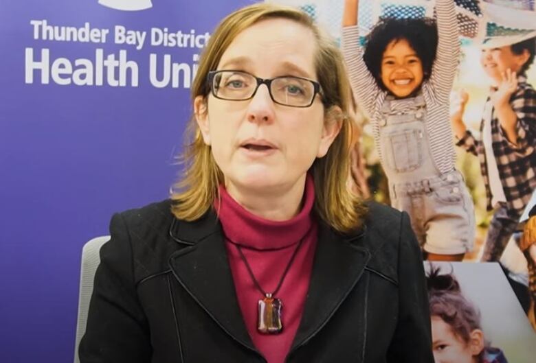 A woman in a black blazer and glasses looks at the camera