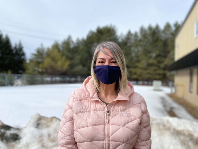 A person wears a dark blue mask and a light pink jacket and stands outside. Behind them is a snowy property.