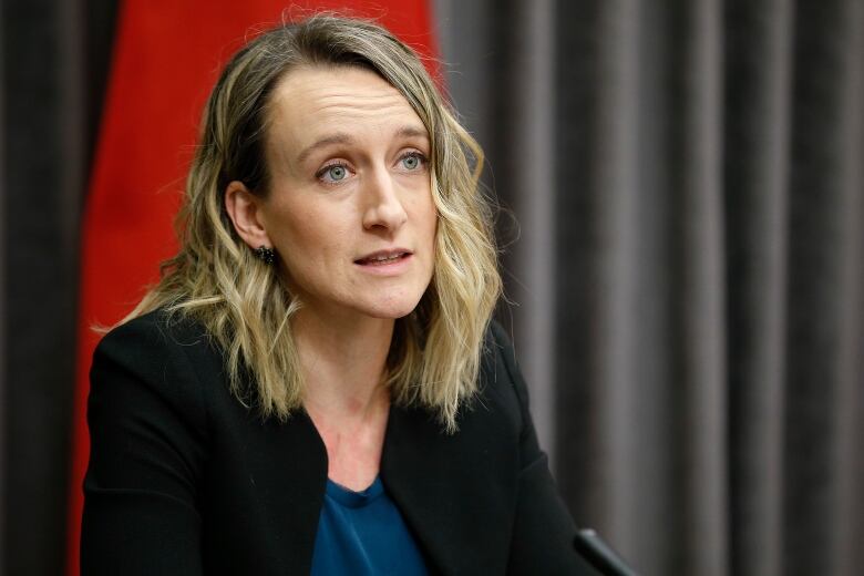 A woman with shoulder-length curled blonde hair is pictured speaking in front of a red flag. She's wearing a black blazer and a turqouise top.