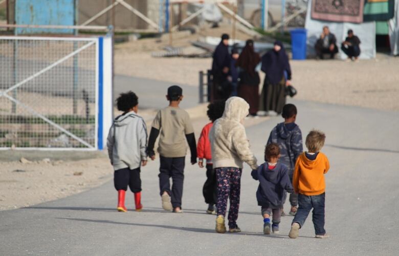 Seven young children, some holding hands, are seen walking along a paved road as women watch in the distance.