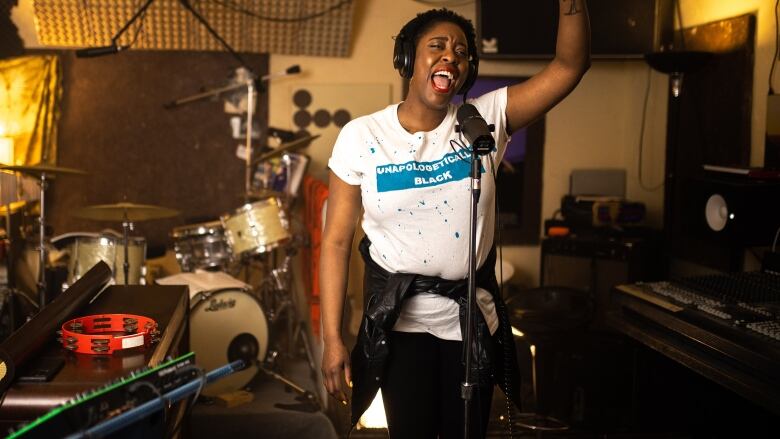 A Black woman wearing a white t-shirt sings at a mic in a music studio