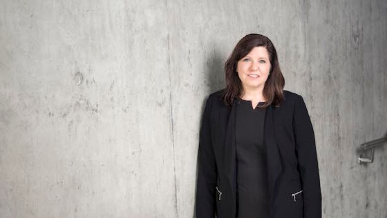 A woman with shoulder-length brown hair, wearing a black jacket and blouse, stands beside a concrete wall and looks into the camera.