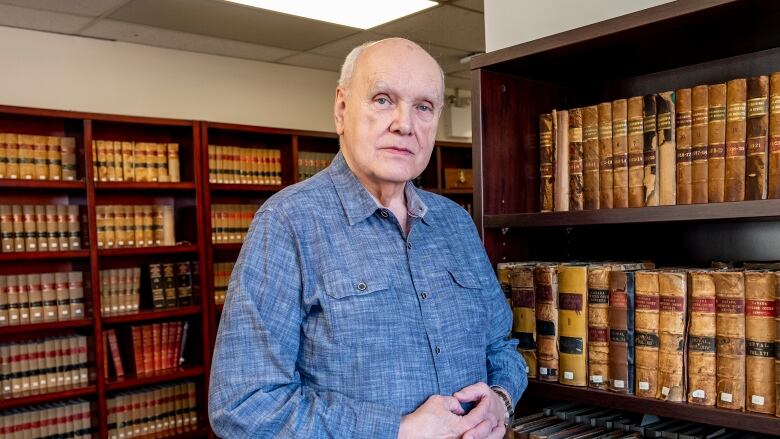 A man wearing a blue button up shirt stands in front of dark wooden third bookshelves that are lined with old books. 