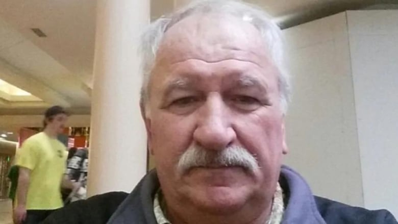 An older man with white hair and a white moustache looks directly at the camera.