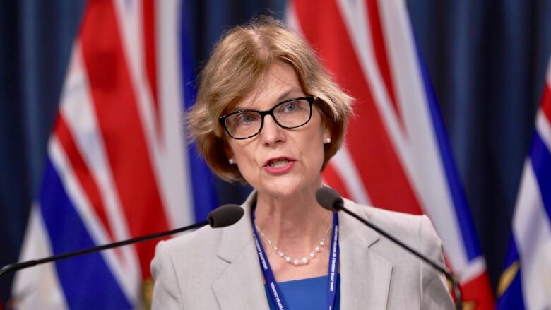 A white woman wearing a grey blazer speaks in front of two B.C. flags.