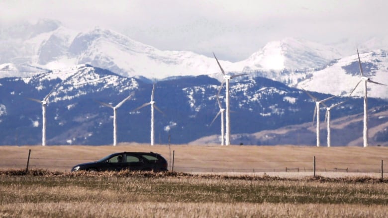 An SUV drives along, with the backdrop of wind turbines in a field, and the snow-peaked Rockies behind.