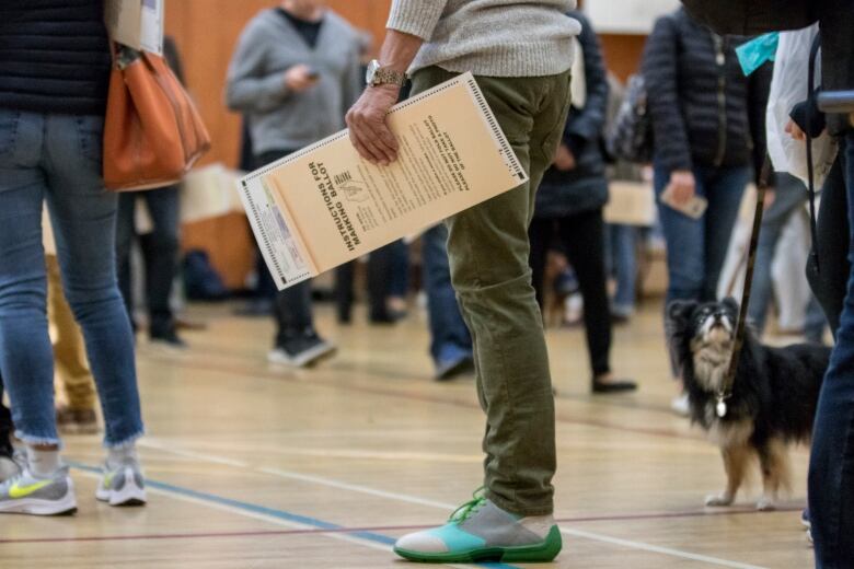 A number of people's legs at a polling station. One person is holding a voter's guide.