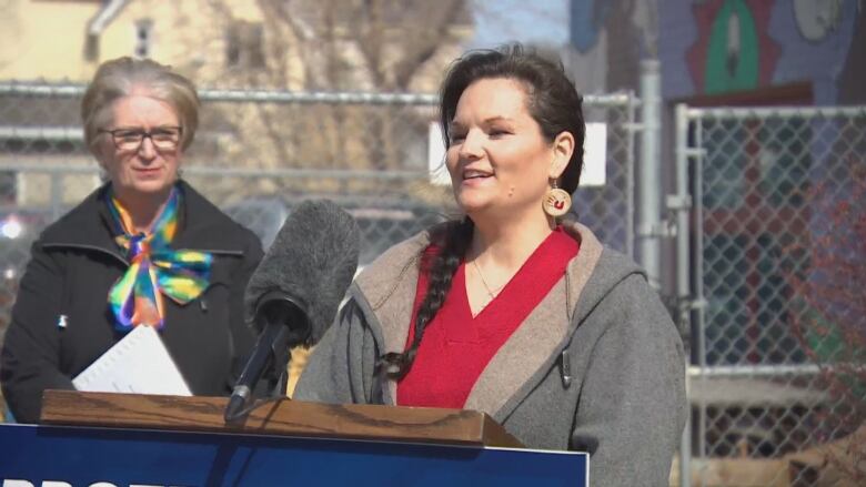 A woman stands in front of a microphone at a podium outside.