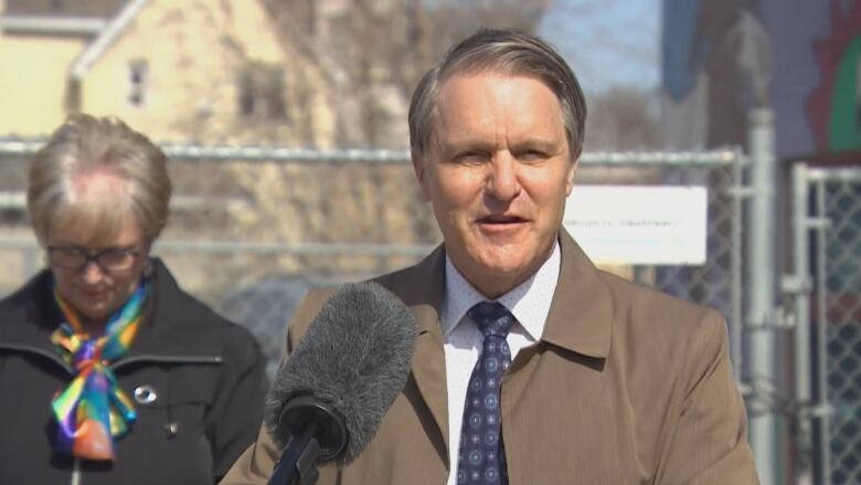 A man in a brown coat speaks at a podium outside.