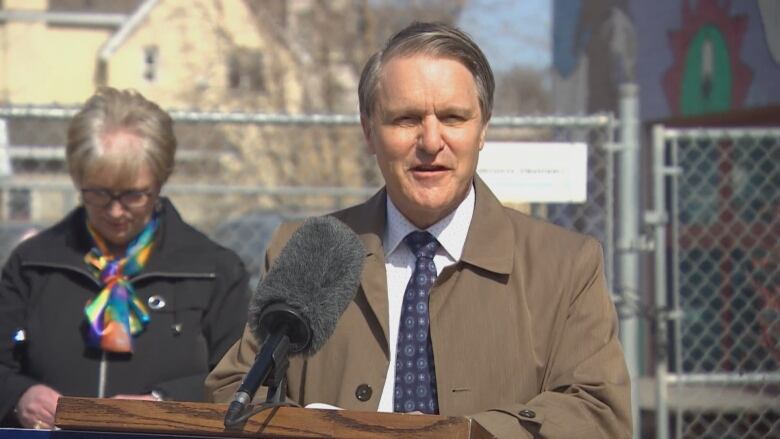 A man in a brown coat speaks at a podium outside.