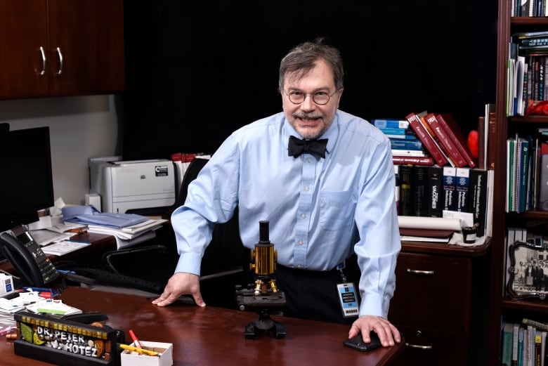 A man with glasses, a blue shirt and a bowtie stands in front of a wooden desk in an office. On the desk is a microscope.