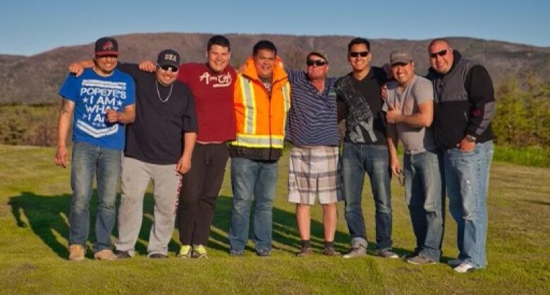 Eight men stand with their arms around each other outside on green grass.