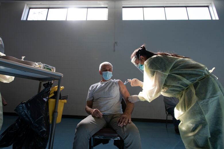 A health-care worker wearing a yellow gown over her clothing, along with a face shield and face mask, administers a vaccine into the arm of an elderly man.