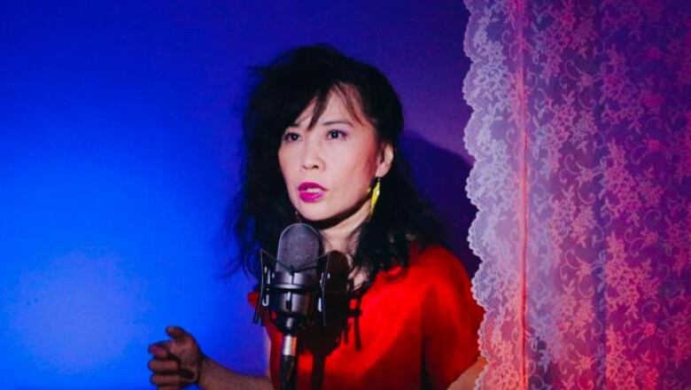 Woman with black hair pulled to one side wearing a red shirt, standing in front of a microphone against a blue backdrop.