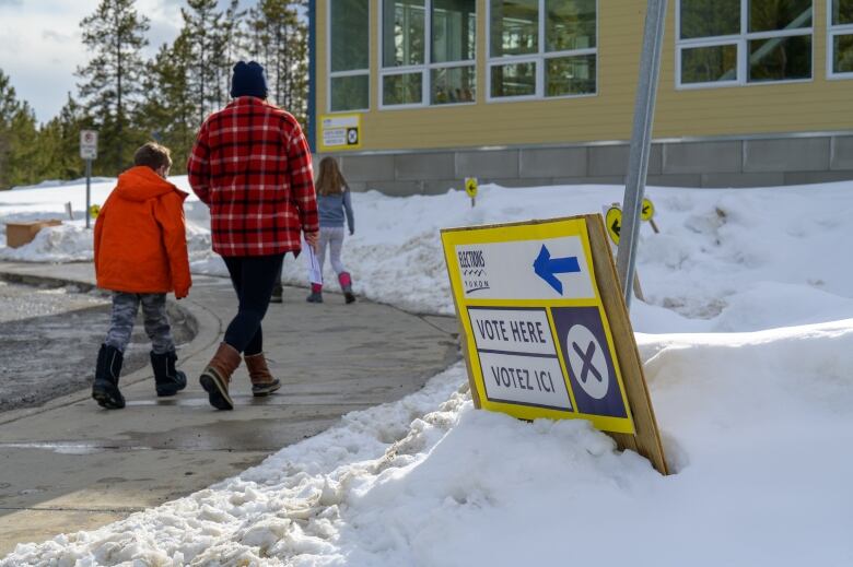 An adult and two children are seen on a snowy day walking toward a building, past a sign reading 'Vote Here.'