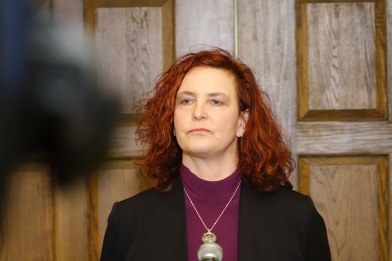 Woman with red hair in purple shirt and black sweater with back to wooden door.