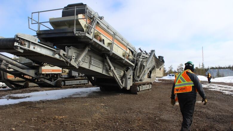 A man in a Vital metals coat walks by a big piece of heavy machinery.