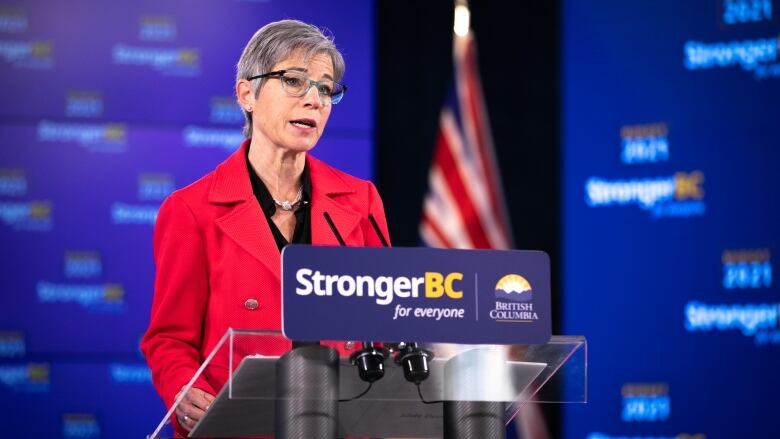 Selina Robinson stands at a podium with 'StrongerBC for Everyone' on it. She is a white woman with blue thin-framed glasses, wearing a red top.