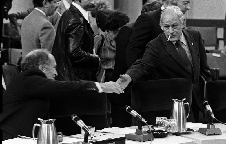 Black-and-white photo of two men reaching across a table to shake hands.