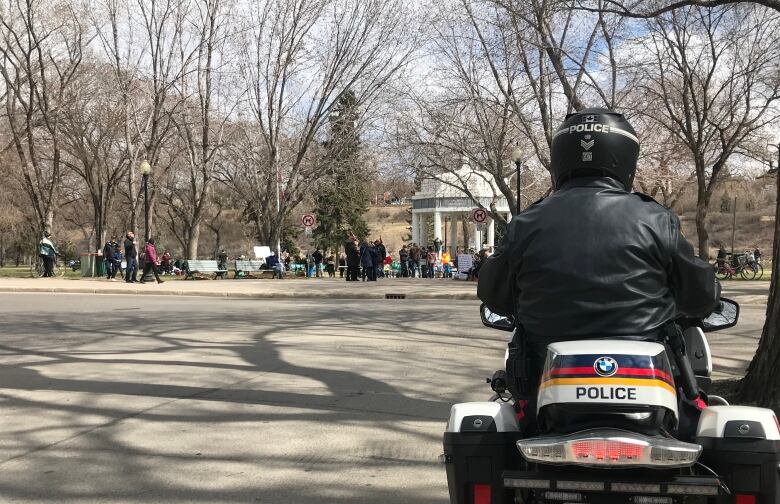 A police officer seated on a police motorcycle watches a large group of people attending an anti-lockdown rally at a sunny park.   