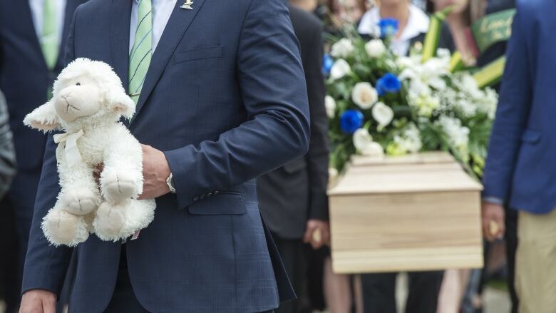 A man holds a stuffed animal. A casket is visible behind him. 