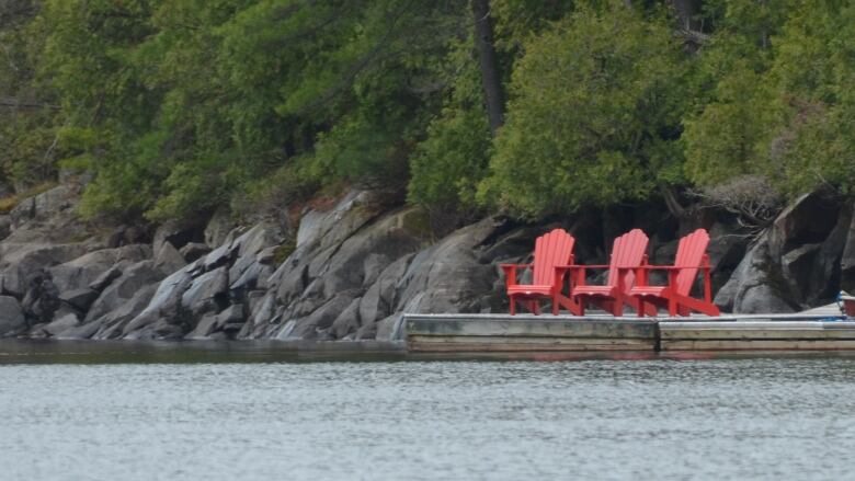 Three red chairs on a dock next a river with trees in the background