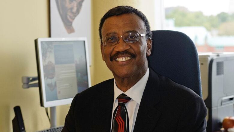 A Black man is seen sitting in an office wearing a black suit