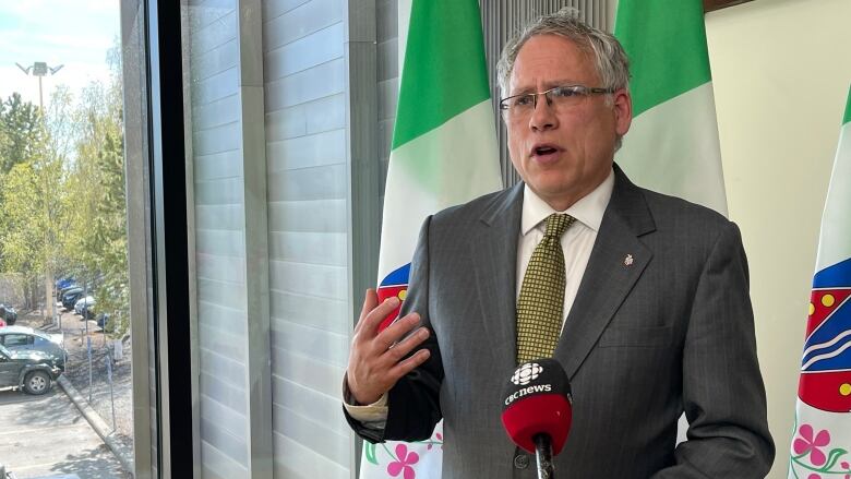 A man in a suit stands at a podium with a CBC microphone and a backdrop of Yukon flags.