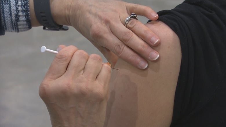 A closeup shows a person getting an injection in their shoulder.