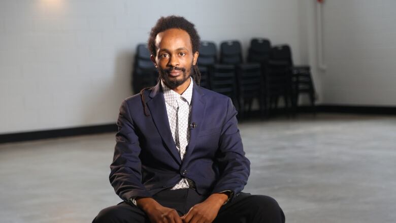 A man wearing a suit sitting in an empty room with grey floors and off-white walls