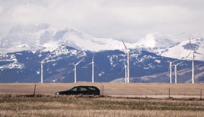 A row of wind turbines is set against a backdrop of mountains. A black car can be seen in the foreground. 