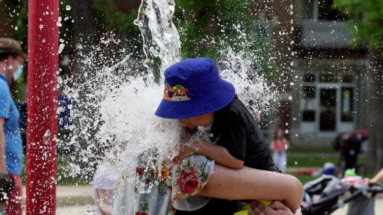 An adult holds a child wearing a bucket hat as they walk under a water sprayer at a splash pad.