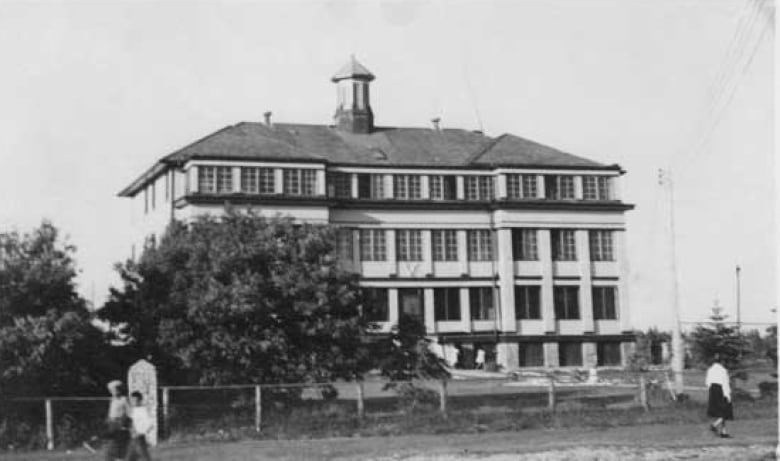 A black and white photo shows people walking past a large, multi-storey building. 