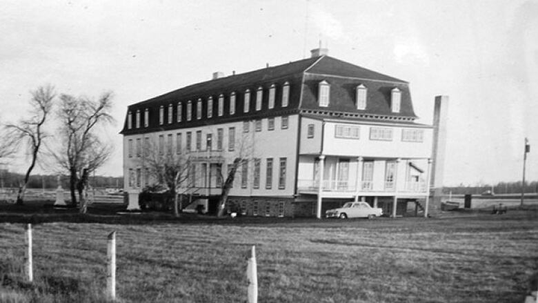 Older black-and-white photo of a building in a field with a few trees and a car out front.