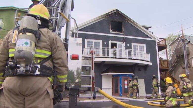 A volunteer firefighter stands with his back turned, facing a large structure that appears to have burned near the roof. Other firefighters are seen to the right.