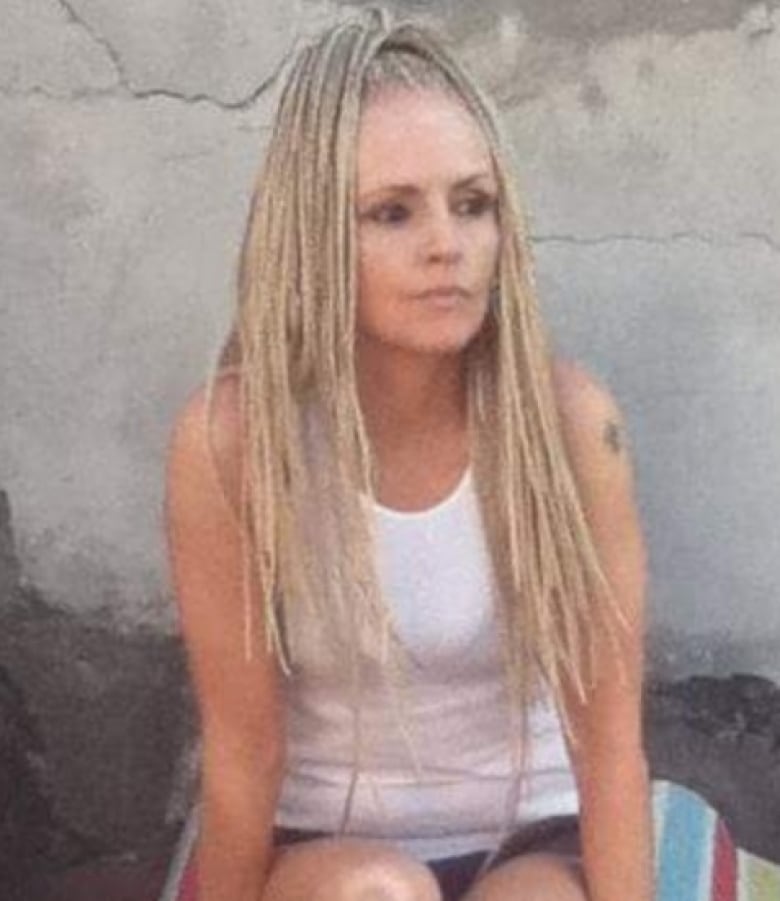 A woman with long blond hair is wearing a white tank top and shorts. She is sitting down and looking into the distance. 