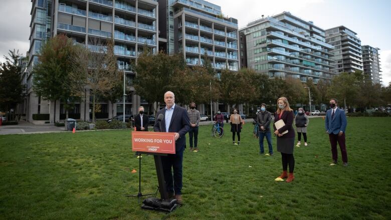 A balding man speaks at an orange podium on a lawn, while people wearing masks stand behind him.