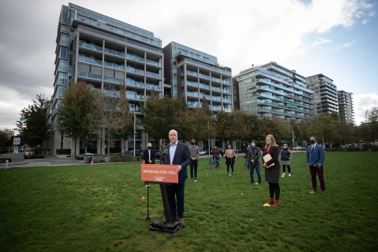 A balding man speaks at an orange podium on a lawn, while people wearing masks stand behind him.