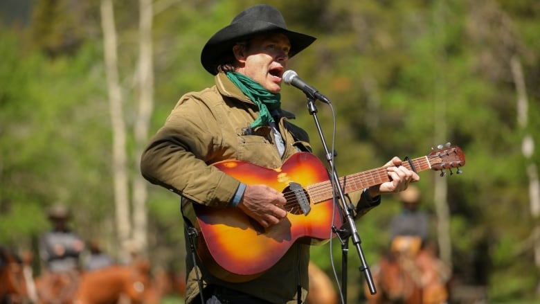 Corb Lund signs into a microphone and plays a guitar in an outdoor setting in front of trees.