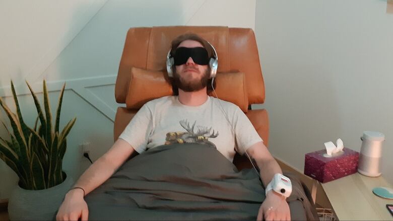 A man wearing an eye mask and headphones sits in a recliner with a monitor on his wrist. 