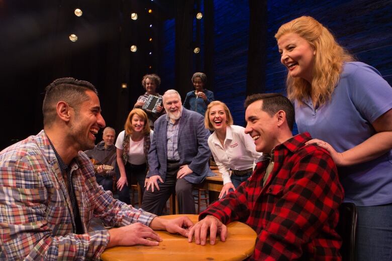 Two people sitting across from each other at a table that's on a theatre stage. Others are watching them and smiling.