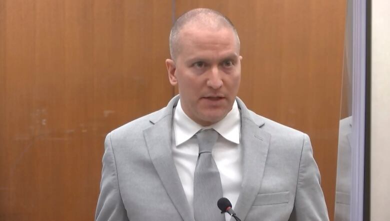 A man in a grey suit and tie addresses a courtroom.